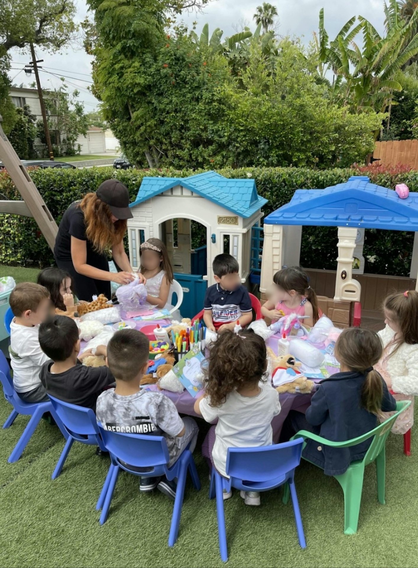 Children gathered around an outdoor craft table with playhouses in the background