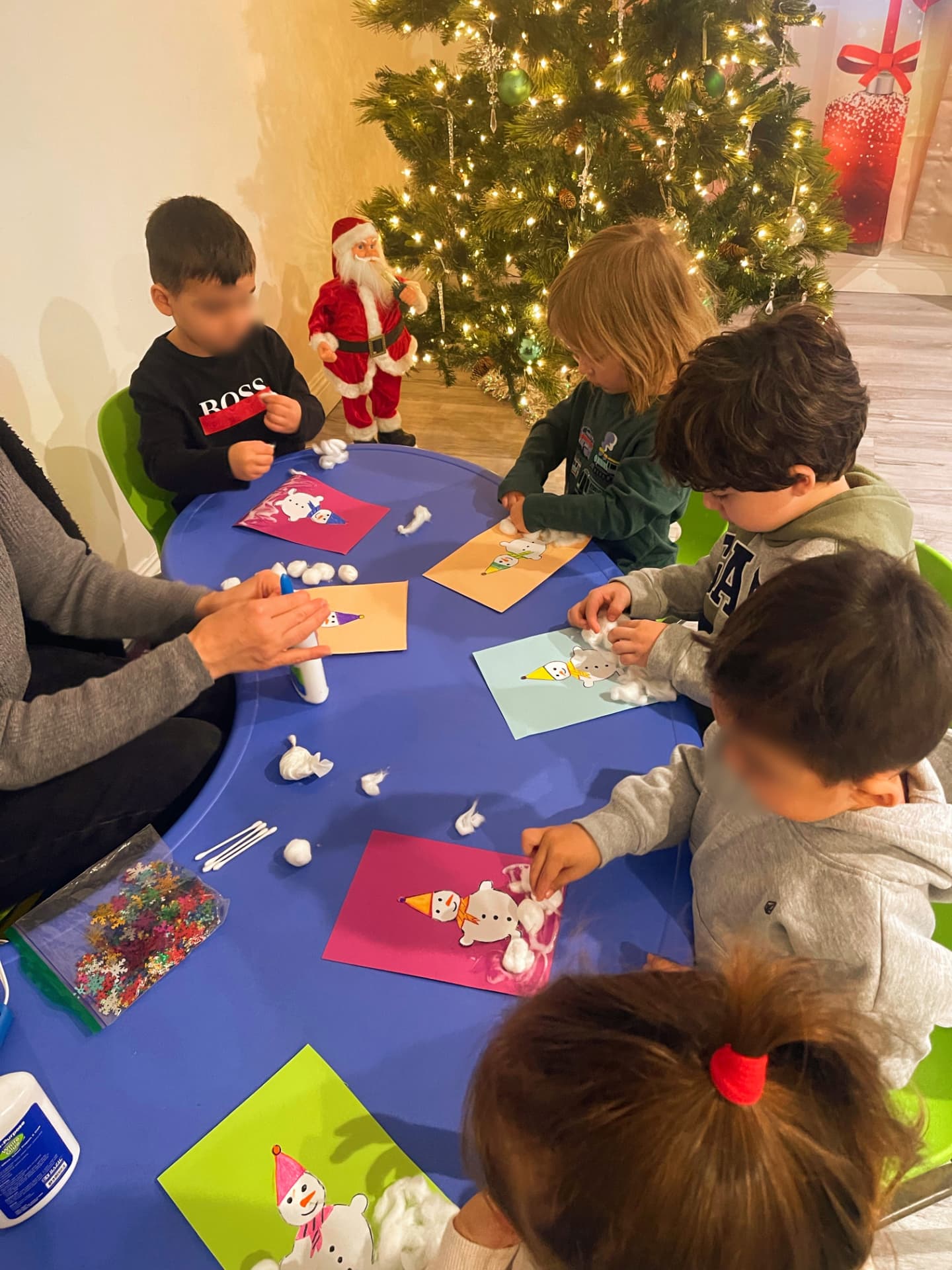 Children making cotton-ball snowman crafts at a table beside a lit Christmas tree