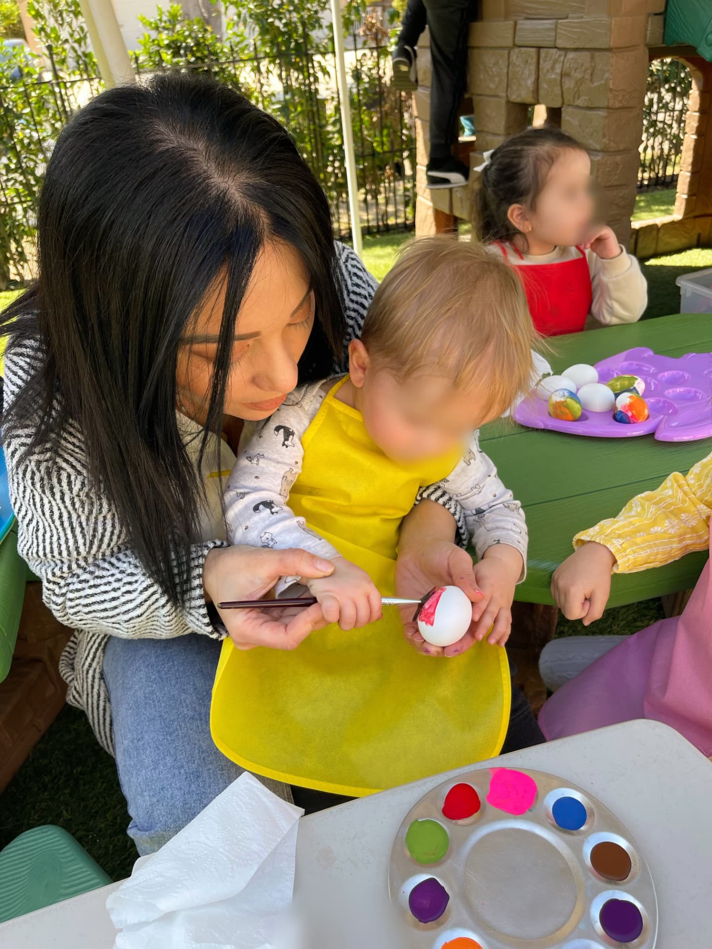 Caregiver helping a toddler paint an egg at an outdoor picnic table
